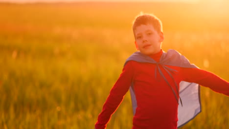 A-boy-dressed-as-a-super-hero-standing-in-a-mask-and-a-red-cloak-runs-laughing-at-the-sunset-in-the-summer-field.-Summer-night-the-boy-dreams-and-heroic-deeds-and-comic-books